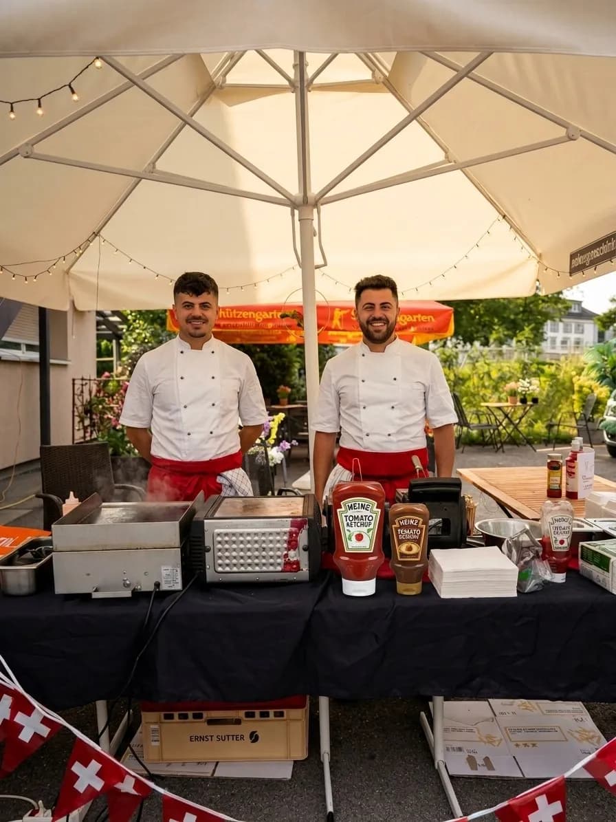 Zwei aus dem Küchenteam an einem Grillstand im Freien unter einem Sonnenschirm
